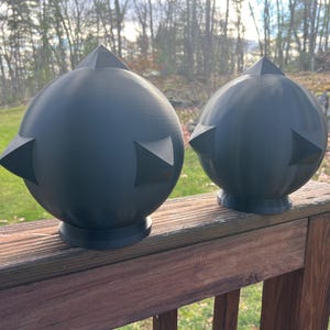 Op de afbeelding: Twee donkergrijze, bolvormige decoratieve objecten met driehoekige punten. De bollen staan op een houten dek, met een natuurlijke achtergrond van bomen en gras. De objecten hebben een modern, geometrisch ontwerp.