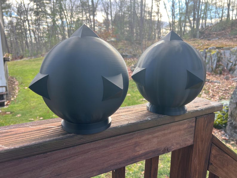 Op de afbeelding: Twee donkergrijze, bolvormige decoratieve objecten met driehoekige punten. De bollen staan op een houten dek, met een natuurlijke achtergrond van bomen en gras. De objecten hebben een modern, geometrisch ontwerp.