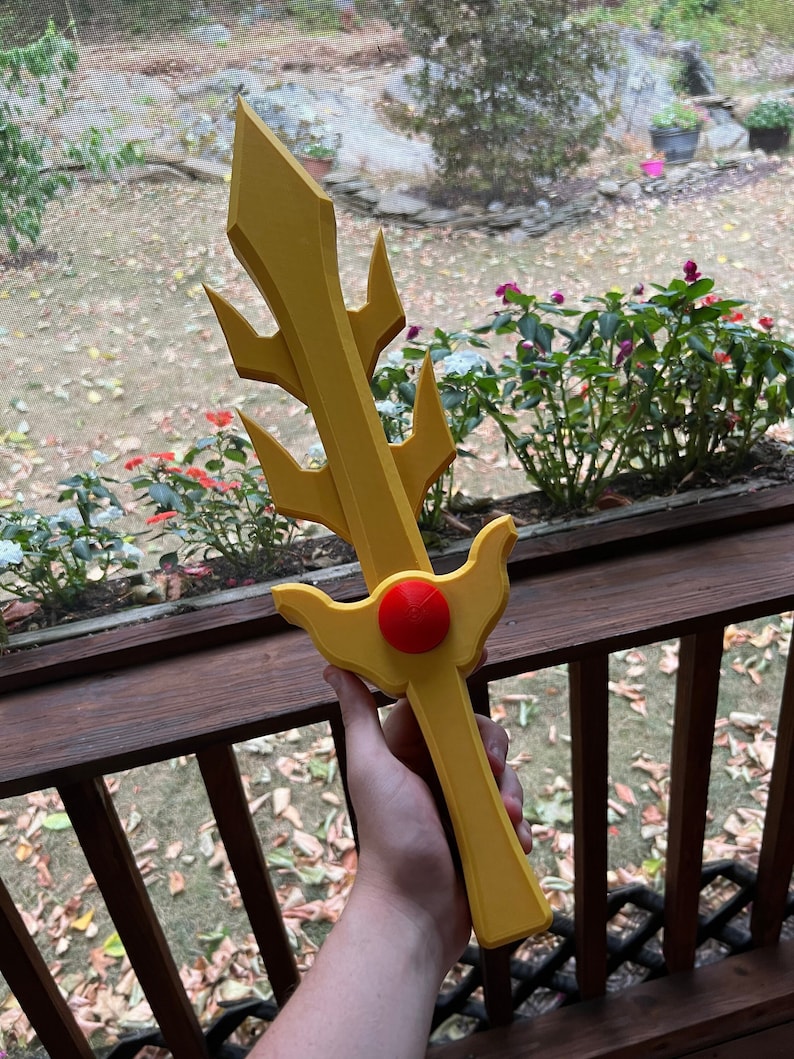 May include: A yellow plastic sword with a red gem in the center of the handle. The sword has a pointed tip and a decorative design with spikes.