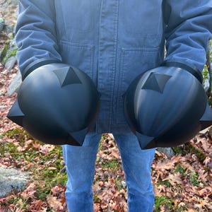 Op de afbeelding: Twee grote, zwarte, bolvormige objecten met driehoekige uitsteeksels en een centraal driehoekig ontwerp. De objecten worden vastgehouden voor een persoon die een blauwe jas en jeans draagt, tegen een achtergrond van gevallen bladeren.
