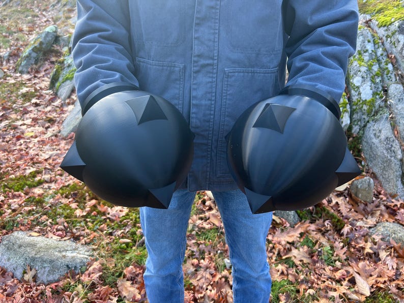 Op de afbeelding: Twee grote, zwarte, bolvormige objecten met driehoekige uitsteeksels en een centraal driehoekig ontwerp. De objecten worden vastgehouden voor een persoon die een blauwe jas en jeans draagt, tegen een achtergrond van gevallen bladeren.