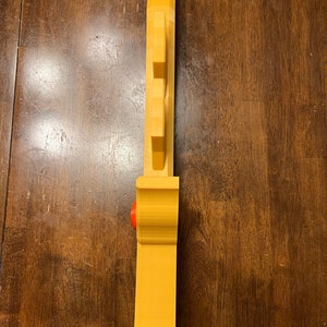 May include: A yellow plastic 3D printed object with a red button on the side. The object has a series of ridges on the front and a pointed top.