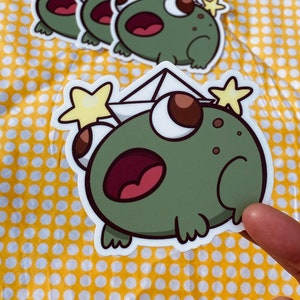 May include: Four green frog stickers with a white envelope and yellow star on top. The frog is wearing a white envelope hat with a yellow star on top. The frog is looking up with its mouth open.