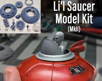 May include: A red and silver model of a flying saucer with a grey alien figure inside. The saucer has a clear dome and a small tail fin. The model is labeled "Li'l Saucer Model Kit (MkII)" and "Updated Dec 2023".