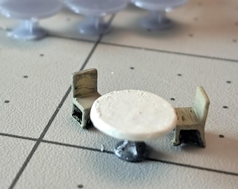 May include: A miniature white table with a round top and a silver base, surrounded by two beige chairs. The table and chairs are made of plastic and are approximately 1 inch tall.