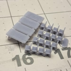 May include: A selection of small, pale grey plastic model components. The parts comprise rectangular pieces, similar to benches, and miniature chairs. The items are arranged on a surface with printed grid lines and the numbers 16 and 17.