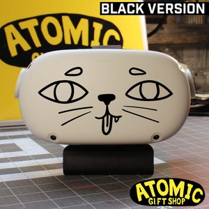May include: A white Oculus Quest 2 virtual reality headset with a black cat face design. The design includes two large eyes, a nose, whiskers, and a tongue sticking out. The headset is on a black stand. The text "ATOMIC GIFT SHOP" is visible in the bottom right corner of the image.