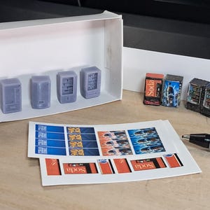 May include: Miniature vending machines in a white box, with printed soda labels and a pen. The vending machines are grey and have detailed designs. The labels are red, blue, and white.