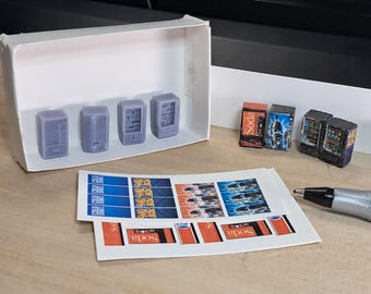 May include: Miniature vending machines in a white box, with printed soda labels and a pen. The vending machines are gray and have detailed designs. The labels are red, blue, and white.