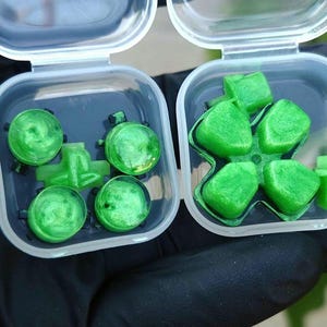 May include: Two clear plastic containers, each open, revealing green objects. One contains four round, green objects and three smaller green pieces. The other holds six green, flower-shaped objects and two small green pieces.