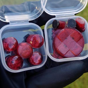 Puede incluir: Dos pequeños recipientes de plástico transparente, cada uno abierto, que revelan tapones rojos y negros. Un recipiente contiene varios tapones redondos, mientras que el otro contiene tapones planos y circulares con un diseño de cruz. Los tapones tienen un aspecto veteado.