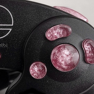 Puede incluir: Un mando negro de Nintendo GameCube con botones frontales rosas. Los botones tienen un diseño de galaxia con remolinos blancos y rosas.
