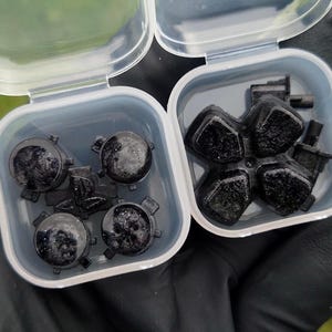May include: Two small, clear plastic containers holding black game controller parts. One container holds four round buttons, and the other holds a directional pad and other components. The parts are dark and appear to be made of plastic.