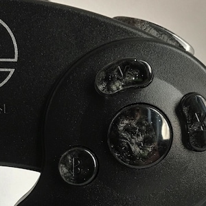 May include: Close-up of a black Nintendo 64 controller with the Super Smash Bros. logo on the top. The controller has a black faceplate with four buttons: A, B, Z, and Start/Pause. The buttons are slightly worn and have a glossy finish.