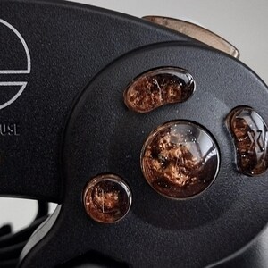 Könnte beinhalten: Nahaufnahme eines schwarzen GameCube-Controllers mit benutzerdefinierten braunen und goldenen Gesichtstasten. Die Tasten haben eine glitzernde, durchscheinende Oberfläche. Der Controller hat ein silbernes Nintendo-Logo und den Text "START/PAUSE" auf der Vorderseite.