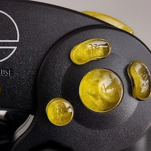 Puede incluir: Un mando negro de Nintendo GameCube con botones frontales amarillos. Los botones están etiquetados como A, B, X, Y y Z. El mando también tiene un botón de inicio/pausa.