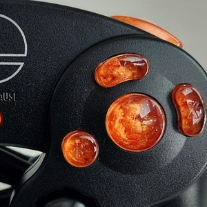 Puede incluir: Primer plano de un mando negro de Nintendo 64 con botones naranjas. Los botones son translúcidos y tienen un acabado brillante. El mando tiene el logotipo de Nintendo y el texto "START/PAUSE".