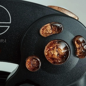 Könnte beinhalten: Ein schwarzer GameCube-Controller mit benutzerdefinierten kupferfarbenen Knöpfen. Die Knöpfe sind durchsichtig und haben eine glitzernde Oberfläche. Der Controller hat die Aufschrift "START/PAUSE" auf der Vorderseite.