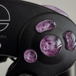 Puede incluir: Un mando de GameCube negro con botones translúcidos de color morado. El mando tiene el logotipo de Nintendo y el texto "START/PAUSE" en la parte delantera.