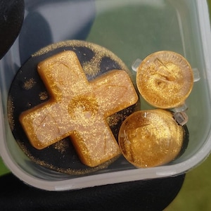 Op de afbeelding: Een doorzichtige plastic container bevat verschillende goudkleurige hars game controller knoppen. De knoppen omvatten een kruisvormig D-pad en twee ronde knoppen. De kruisknop staat op een zwarte achtergrond.