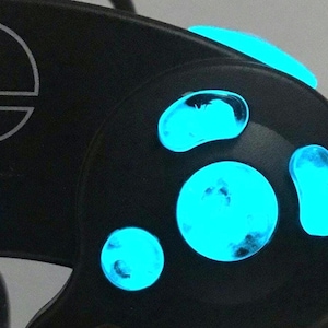 Könnte beinhalten: Nahaufnahme eines schwarzen Videospiel-Controllers mit leuchtend blauen Knöpfen und einem weißen Logo. Der Controller hat ein kreisförmiges Design mit mehreren Knöpfen und einem Steuerkreuz. Die Knöpfe und das Logo leuchten hellblau.
