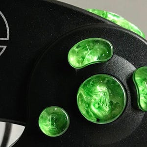 May include: Close-up of a black video game controller with green translucent buttons. The buttons are labelled "A", "B", "X", and "Y". The controller also has a "Start/Pause" button.