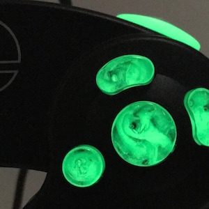 Könnte beinhalten: Ein schwarzer GameCube-Controller mit im Dunkeln leuchtenden Knöpfen. Die Knöpfe sind in Form eines Kreises, Ovals und zweier Bohnen geformt. Die Knöpfe leuchten grün.