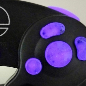 Puede incluir: Un mando de GameCube negro con botones morados. El mando tiene el logotipo de Nintendo en la parte delantera.