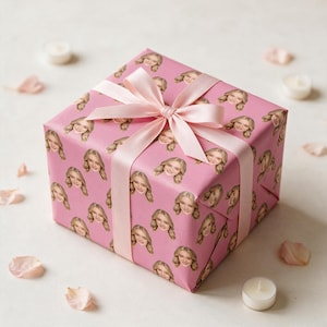 May include: A square gift box wrapped in pink paper featuring a repeating pattern of a smiling person's face. The box is tied with a smooth, light pink satin ribbon arranged in a decorative bow on the top center of the package.