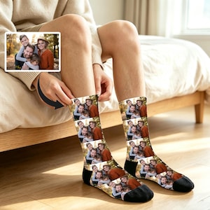 May include: A pair of crew-length socks featuring a repeating photo print of a family of four. The socks have black toe and heel sections. The printed image shows two adults and two children posing outdoors in a wooded area with autumn foliage.