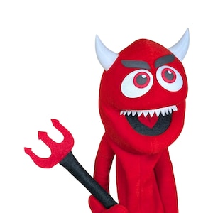 Puede incluir: Una marioneta de mano roja que representa un personaje de diablo con dos cuernos blancos, ojos grandes blancos con pupilas rojas y una boca ancha con dientes blancos afilados. La marioneta sostiene un tridente negro con cabeza roja.