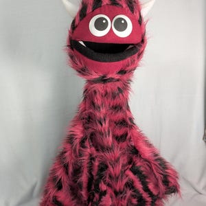 Könnte beinhalten: Eine Handpuppe in Form eines langhalsigen Monsters mit flauschigem, pink-schwarzem Fell. Die Figur hat zwei weiße Hörner, große runde weiße Augen mit schwarzen Pupillen und ein weit geöffnetes Maul mit zwei sichtbaren weißen Fangzähnen.