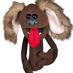 Puede incluir: Una marioneta de mano de forro polar marrón con forma de perro, con largas orejas de piel sintética, grandes ojos de plástico negro, una nariz gris oscuro y una lengua de fieltro rojo brillante que sobresale de una boca abierta.