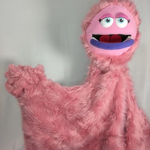 Puede incluir: Una marioneta de mano de peluche rosa con cara redonda, grandes ojos azules, largas pestañas negras y párpados rosas. La marioneta tiene la boca muy abierta con un labio inferior morado, un interior oscuro y una lengua rosa, cubierta de pelo largo.