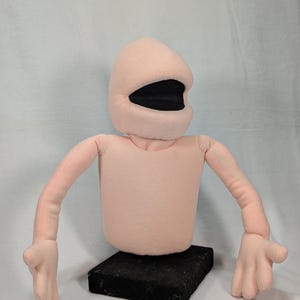 Puede incluir: Un torso de marioneta de felpa rosa claro con cabeza redondeada, una amplia abertura bucal negra y dos brazos largos con manos de cuatro dedos. La marioneta está montada sobre una base rectangular negra e incluye dos varillas de control.