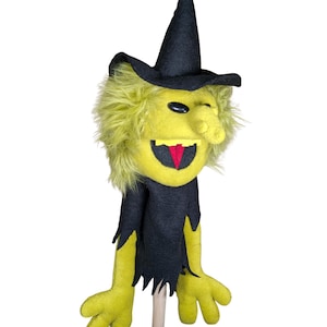 May include: A hand puppet featuring a yellow face with shaggy yellow hair, a black pointed hat, and a black tattered garment. The puppet has a large nose, a wide mouth with yellow teeth, a red tongue, and a single visible black oval eye.