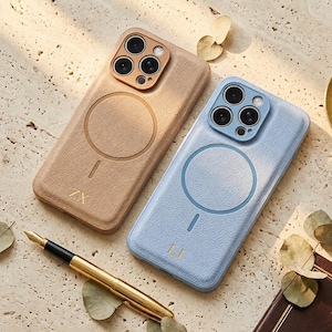 May include: Two phone cases, one tan and one light blue, with a circular design and camera cutouts. The tan case has the initials "ZX" and the blue case has "LJ". A gold fountain pen and dried leaves are also present.
