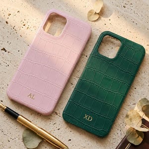 以下が含まれることがあります： クロコダイル柄の2つの携帯電話ケース。1つは金色のイニシャル「AL」が入った薄いピンク色、もう1つはイニシャル「XD」が入った濃い緑色です。金の万年筆と真鍮の皿も画像にあります。