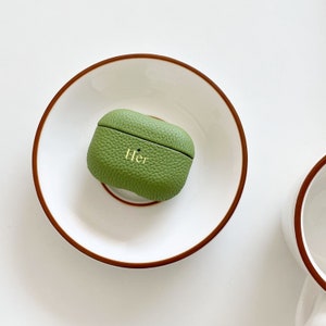 May include: A green leather case for wireless earbuds with the word "Her" embossed in gold lettering. The case is sitting on a white plate with a brown rim.