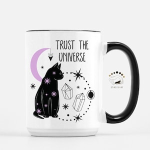 May include: White ceramic mug with a black handle. The mug features a black cat sitting next to a crescent moon with a pink interior. The text "TRUST THE UNIVERSE" is printed in black above the cat. The design also includes stars, crystals, and a circle of dots.