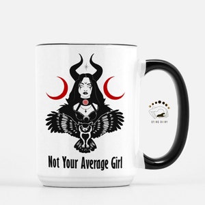 Può includere: Tazza in ceramica bianca con bordo e manico neri. La tazza presenta un'illustrazione in bianco e nero di una donna con corna, lune crescenti e un gufo. Il testo "Not Your Average Girl" è stampato sul davanti.