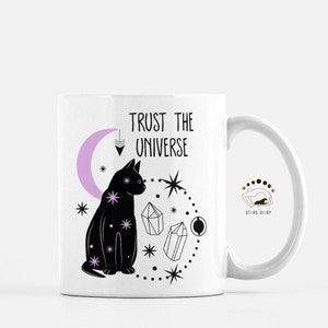 May include: White ceramic mug with a black cat, pink crescent moon, and crystals. The text "TRUST THE UNIVERSE" is printed on the mug. The bottom of the mug has the text "STARS DIARY".