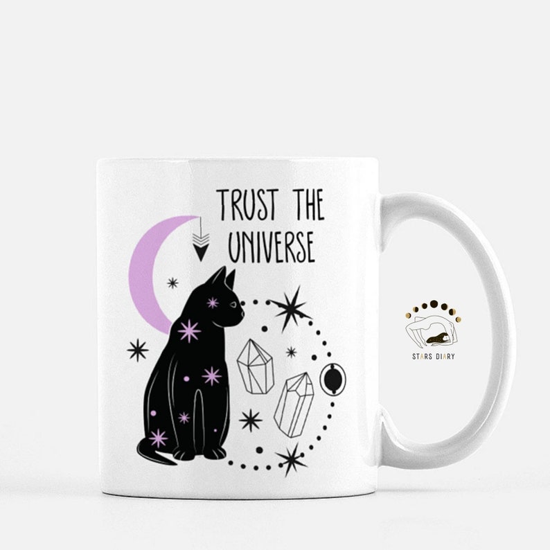 May include: White ceramic mug with a black cat, pink crescent moon, and crystals. The text "TRUST THE UNIVERSE" is printed on the mug. The bottom of the mug has the text "STARS DIARY".