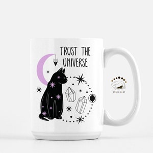 May include: White ceramic mug with a black cat, crystals, and stars design. The text "Trust the Universe" is printed on the mug. The mug has a black handle.