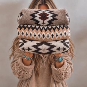 Op de afbeelding: Een stapel van drie gevouwen truien met geometrische patronen in tinten bruin, crème en zwart. De bovenste trui heeft een groot ruitmotief, de middelste een zigzagband en de onderste een herhalend hoekig ontwerp.