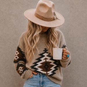 Puede incluir: Un sombrero fedora de fieltro beige con una banda texturizada y una pequeña pluma, combinado con un suéter beige de manga larga con un patrón geométrico en marrón oscuro, negro y blanco, jeans azul claro desgastados y un anillo de turquesa.