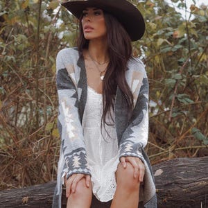 May include: A woman wearing a brown cowboy hat, a grey patterned cardigan, a white dress, and brown cowboy boots. She is sitting on a log in a wooded area, surrounded by foliage and fallen leaves.