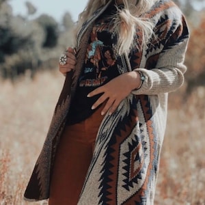 May include: A long, open-front cardigan in a neutral color with a Southwestern-inspired geometric pattern in shades of brown, navy, and cream. The cardigan is paired with a black graphic tee and rust-colored pants. A silver ring and bracelet are also visible.