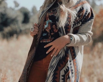 May include: A long, open-front cardigan in a neutral color with a Southwestern-inspired geometric pattern in shades of brown, navy, and cream. The cardigan is paired with a black graphic tee and rust-colored pants. A silver ring and bracelet are also visible.