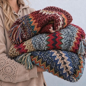 May include: A thick, folded knit blanket featuring a repeating chevron pattern in deep red, navy blue, mustard yellow, and cream. The textile has a textured, chunky yarn construction and is finished with multi-colored fringe tassels on the edges.